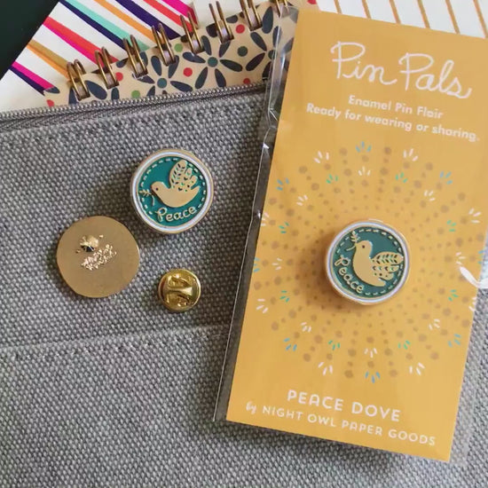 Front of enamel pin is blue with gold dove and Peace text and gold accents. Back of pin is gold with the Night Owl Paper Goods script logo. Pin is mounted on a backer card that says "Pin Pals - Enamel Pin Flair - Ready for wearing or sharing - PEACE DOVE - by Night Owl Paper Goods" and is packaged in a clear bag.