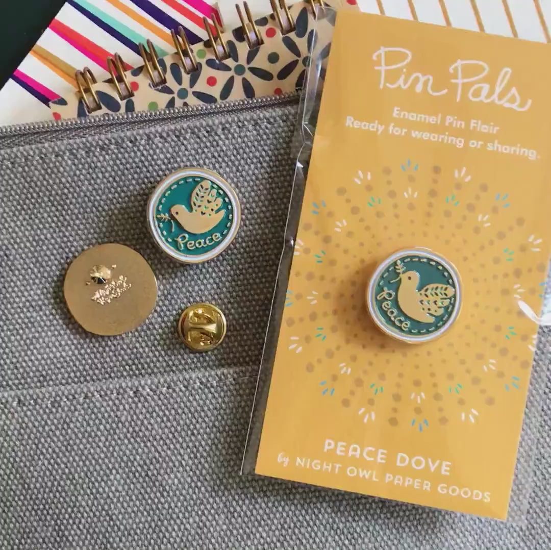 Front of enamel pin is blue with gold dove and Peace text and gold accents. Back of pin is gold with the Night Owl Paper Goods script logo. Pin is mounted on a backer card that says "Pin Pals - Enamel Pin Flair - Ready for wearing or sharing - PEACE DOVE - by Night Owl Paper Goods" and is packaged in a clear bag.