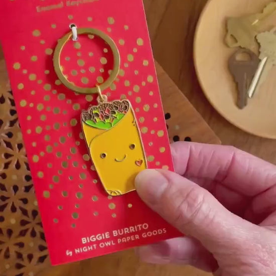 Front of enamel keychain is a smiling burrito with gold accents. Keychain is mounted on a backer card that says "Key Charm - Enamel Keychain - BIGGIE BURRITO by Night Owl Paper Goods" and is packaged in a clear box.