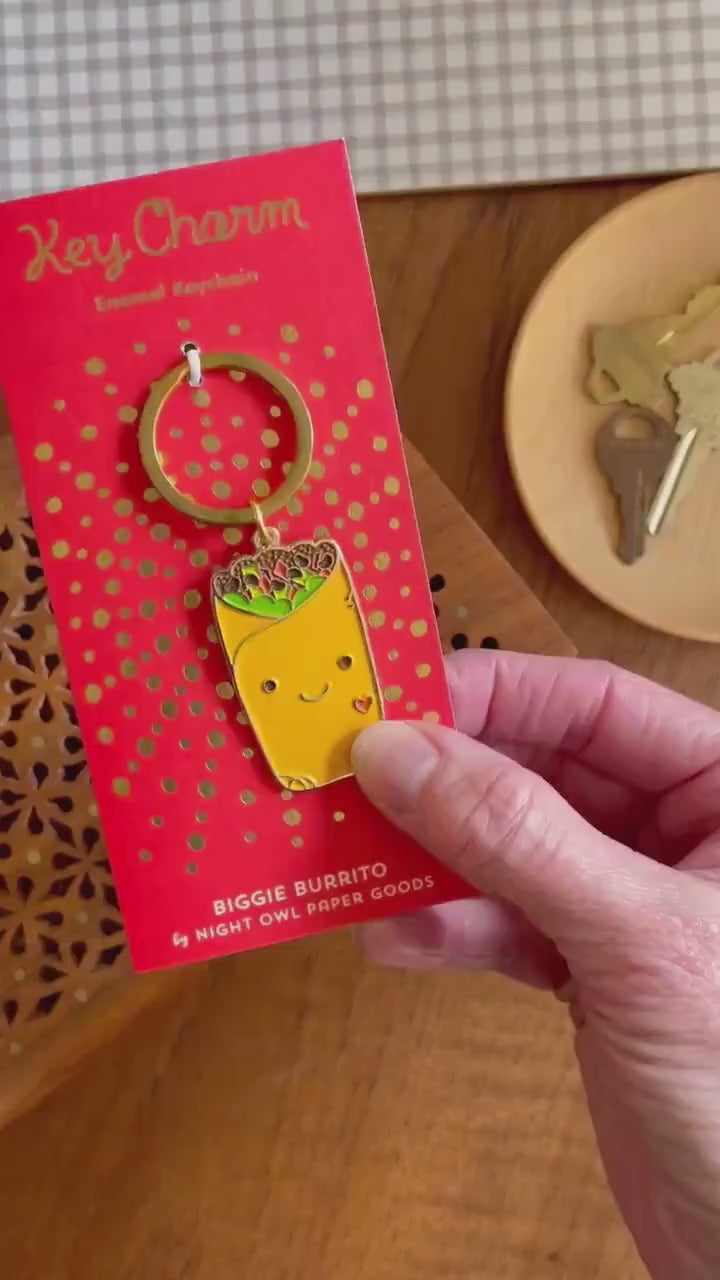 Front of enamel keychain is a smiling burrito with gold accents. Keychain is mounted on a backer card that says "Key Charm - Enamel Keychain - BIGGIE BURRITO by Night Owl Paper Goods" and is packaged in a clear box.
