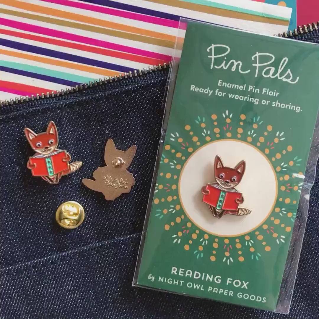 Front of enamel pin is a fox reading a book with gold accents and a small white heart. Back of pin is gold with the Night Owl Paper Goods script logo. Pin is mounted on a backer card that says "Pin Pals - Enamel Pin Flair - Ready for wearing or sharing - READING FOX - by Night Owl Paper Goods" and is packaged in a clear bag.