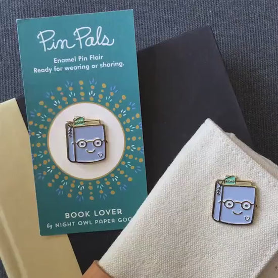 Front of enamel pin is a smiling book wearing glasses with gold accents. Back of pin is gold with the Night Owl Paper Goods script logo. Pin is mounted on a backer card that says "Pin Pals - Enamel Pin Flair - Ready for wearing or sharing - BOOK LOVER - by Night Owl Paper Goods" and is packaged in a clear bag.