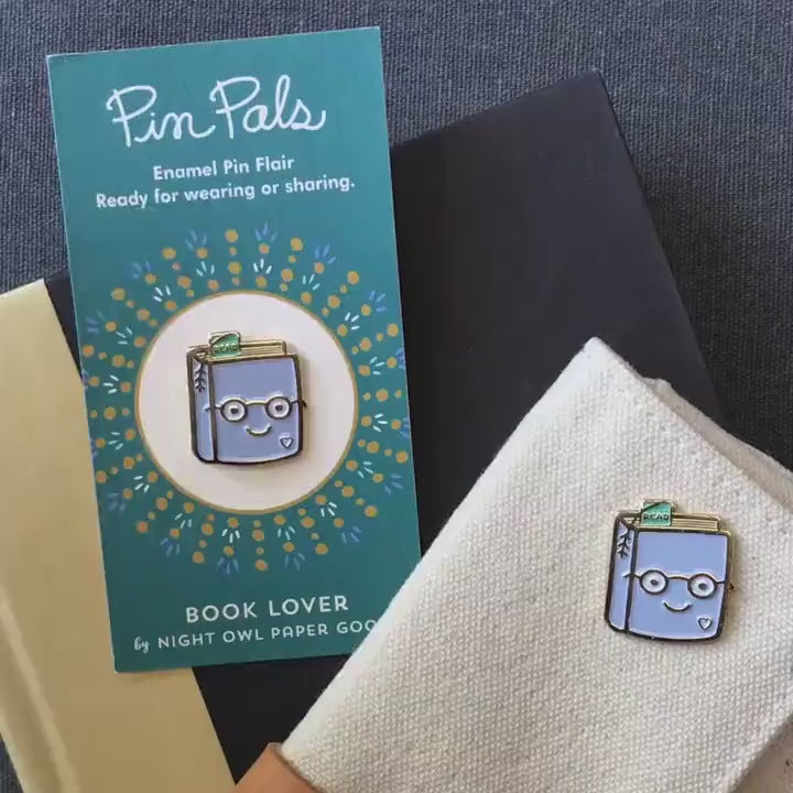 Front of enamel pin is a smiling book wearing glasses with gold accents. Back of pin is gold with the Night Owl Paper Goods script logo. Pin is mounted on a backer card that says "Pin Pals - Enamel Pin Flair - Ready for wearing or sharing - BOOK LOVER - by Night Owl Paper Goods" and is packaged in a clear bag.
