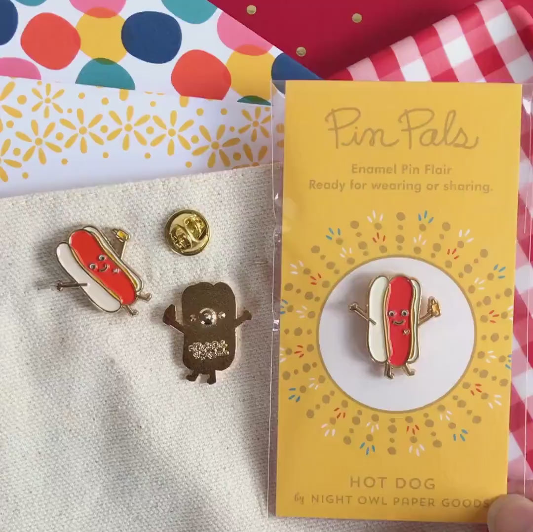 Front of enamel pin is smiling hot dog with gold accents and a small white heart. Back of pin is gold with the Night Owl Paper Goods script logo. Pin is mounted on a backer card that says "Pin Pals - Enamel Pin Flair - Ready for wearing or sharing - HOT DOG - by Night Owl Paper Goods" and is packaged in a clear bag.