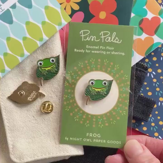 Front of enamel pin is a green frog on top of a lilypad with gold accents. Back of pin is gold with the Night Owl Paper Goods script logo. Pin is mounted on a backer card that says "Pin Pals - Enamel Pin Flair - Ready for wearing or sharing - FROG - by Night Owl Paper Goods" and is packaged in a clear bag.