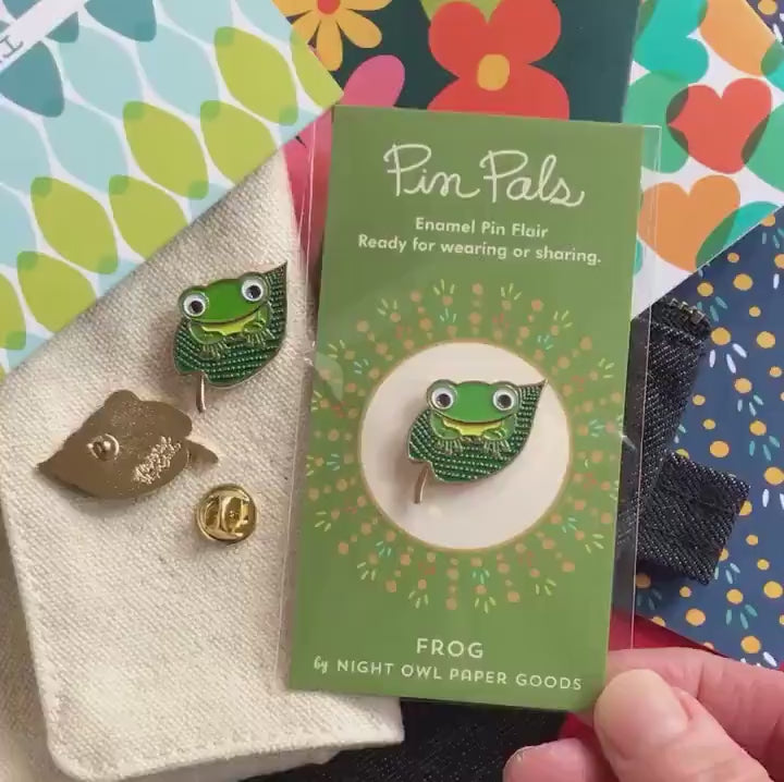 Front of enamel pin is a green frog on top of a lilypad with gold accents. Back of pin is gold with the Night Owl Paper Goods script logo. Pin is mounted on a backer card that says "Pin Pals - Enamel Pin Flair - Ready for wearing or sharing - FROG - by Night Owl Paper Goods" and is packaged in a clear bag.