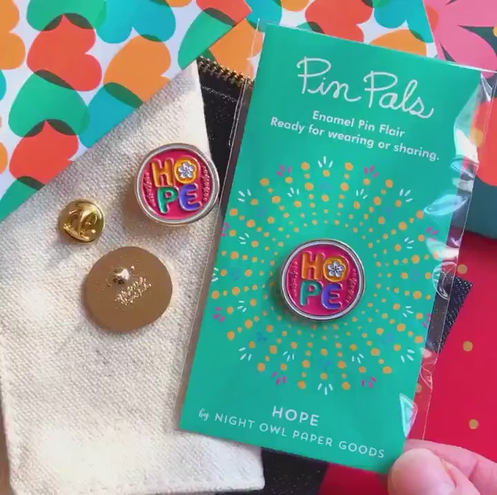 Front of enamel pin is pink with colorful HOPE text, a small flower in the middle of the "O" and gold accents. Back of pin is gold with the Night Owl Paper Goods script logo. Pin is mounted on a backer card that says "Pin Pals - Enamel Pin Flair - Ready for wearing or sharing - HOPE - by Night Owl Paper Goods" and is packaged in a clear bag.