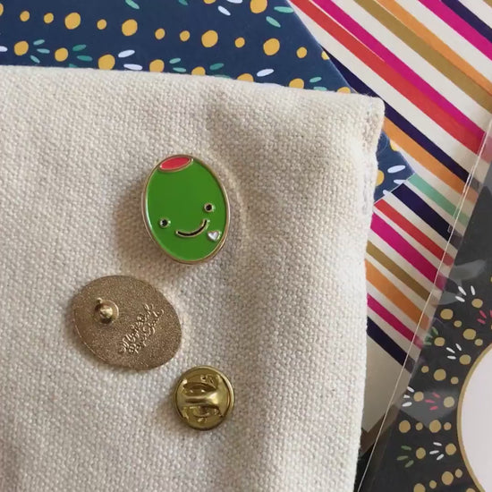 Front of enamel pin is smiling green olive with gold accents and a small white heart. Back of pin is gold with the Night Owl Paper Goods script logo. Pin is mounted on a backer card that says "Pin Pals - Enamel Pin Flair - Ready for wearing or sharing - OLIVE LOVE - by Night Owl Paper Goods" and is packaged in a clear bag.