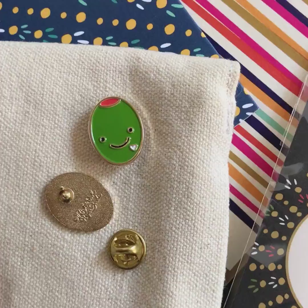 Front of enamel pin is smiling green olive with gold accents and a small white heart. Back of pin is gold with the Night Owl Paper Goods script logo. Pin is mounted on a backer card that says "Pin Pals - Enamel Pin Flair - Ready for wearing or sharing - OLIVE LOVE - by Night Owl Paper Goods" and is packaged in a clear bag.