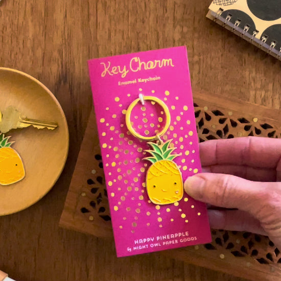 Front of enamel keychain is a smiling pineapple with gold accents. Keychain is mounted on a backer card that says "Key Charm - Enamel Keychain - HAPPY PINEAPPLE by Night Owl Paper Goods" and is packaged in a clear box.