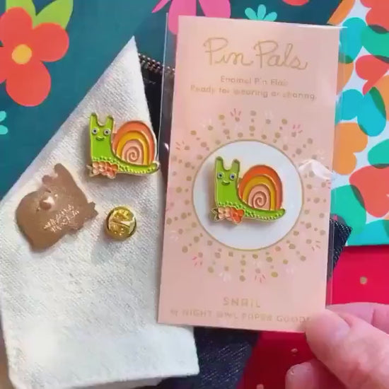 Front of enamel pin is a smiling snail with gold accents. Back of pin is gold with the Night Owl Paper Goods script logo. Pin is mounted on a backer card that says "Pin Pals - Enamel Pin Flair - Ready for wearing or sharing - SNAIL - by Night Owl Paper Goods" and is packaged in a clear bag.
