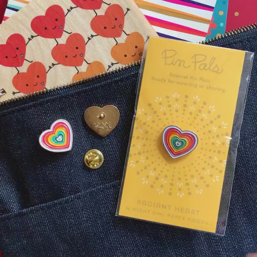 Front of enamel pin is a heart with radiant rainbow colors and gold accents. Back of pin is gold with the Night Owl Paper Goods script logo. Pin is mounted on a backer card that says "Pin Pals - Enamel Pin Flair - Ready for wearing or sharing - RADIANT HEART - by Night Owl Paper Goods" and is packaged in a clear bag.