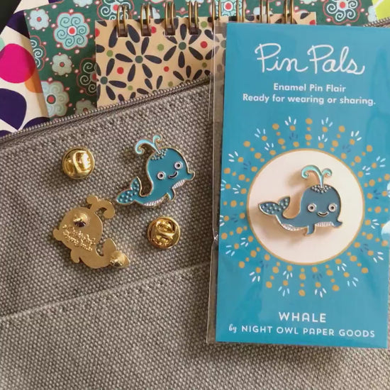 Front of enamel pin is a smiling blue whale with gold accents. Back of pin is gold with the Night Owl Paper Goods script logo. Pin is mounted on a backer card that says "Pin Pals - Enamel Pin Flair - Ready for wearing or sharing - WHALE - by Night Owl Paper Goods" and is packaged in a clear bag.