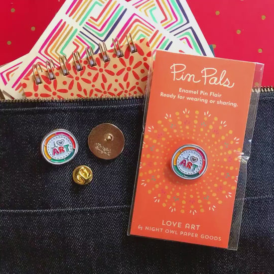 Front of enamel pin is a color wheel with "I heart art" text and gold accents. Back of pin is gold with the Night Owl Paper Goods script logo. Pin is mounted on a backer card that says "Pin Pals - Enamel Pin Flair - Ready for wearing or sharing - LOVE ART - by Night Owl Paper Goods" and is packaged in a clear bag.