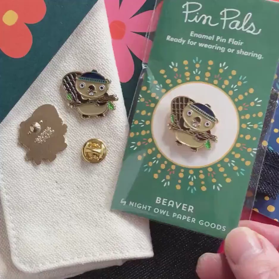 Front of enamel pin is a smiling beaver holding logs with gold accents. Back of pin is gold with the Night Owl Paper Goods script logo. Pin is mounted on a backer card that says "Pin Pals - Enamel Pin Flair - Ready for wearing or sharing - BEAVER - by Night Owl Paper Goods" and is packaged in a clear bag.