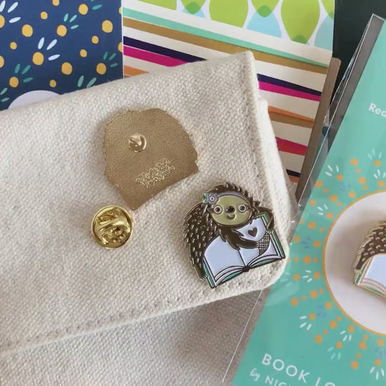 Front of enamel pin is a cute sloth reading a book with gold accents. Back of pin is gold with the Night Owl Paper Goods script logo. Pin is mounted on a backer card that says "Pin Pals - Enamel Pin Flair - Ready for wearing or sharing - BOOK LOVER SLOTH - by Night Owl Paper Goods" and is packaged in a clear bag.