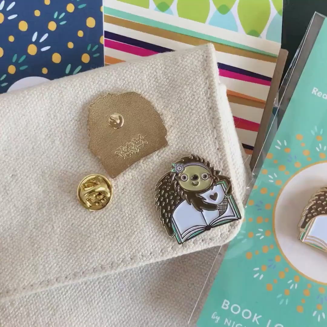 Front of enamel pin is a cute sloth reading a book with gold accents. Back of pin is gold with the Night Owl Paper Goods script logo. Pin is mounted on a backer card that says "Pin Pals - Enamel Pin Flair - Ready for wearing or sharing - BOOK LOVER SLOTH - by Night Owl Paper Goods" and is packaged in a clear bag.