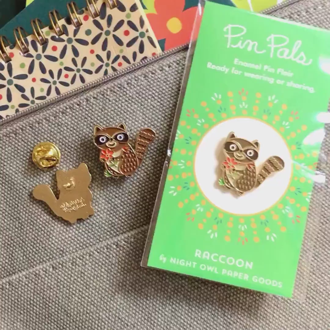 Front of enamel pin is a smiling raccoon holding a red flower with gold accents. Back of pin is gold with the Night Owl Paper Goods script logo. Pin is mounted on a backer card that says "Pin Pals - Enamel Pin Flair - Ready for wearing or sharing - RACCOON - by Night Owl Paper Goods" and is packaged in a clear bag.