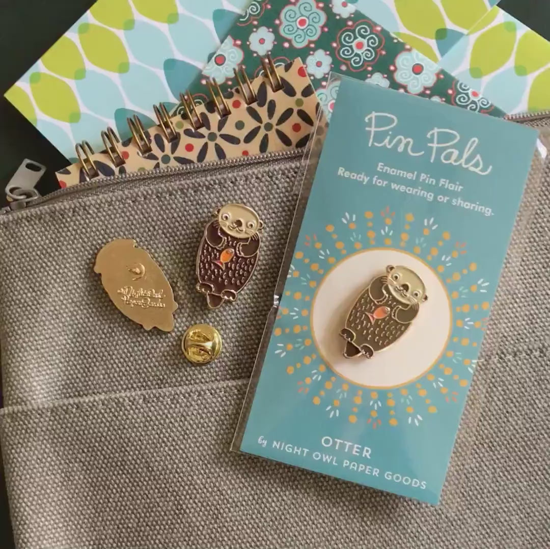 Front of enamel pin is a brown river otter with gold accents and an orange fish on its belly. Back of pin is gold with the Night Owl Paper Goods script logo. Pin is mounted on a backer card that says "Pin Pals - Enamel Pin Flair - Ready for wearing or sharing - OTTER - by Night Owl Paper Goods" and is packaged in a clear bag.