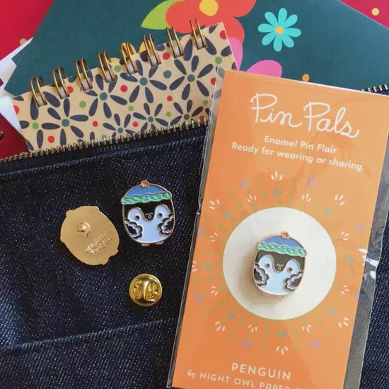 Front of enamel pin is a penguin wearing a stocking cap with gold accents. Back of pin is gold with the Night Owl Paper Goods script logo. Pin is mounted on a backer card that says "Pin Pals - Enamel Pin Flair - Ready for wearing or sharing - PENGUIN - by Night Owl Paper Goods" and is packaged in a clear bag.