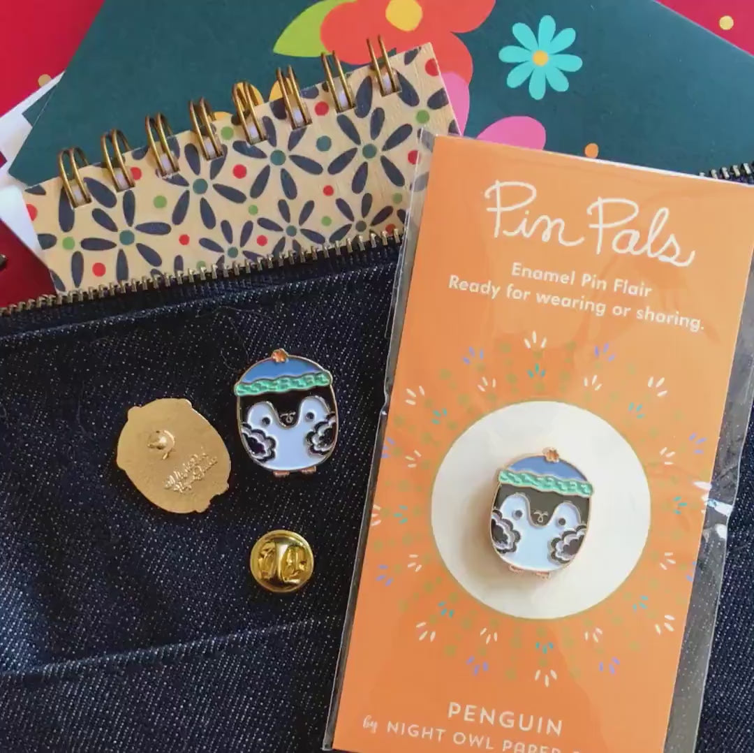 Front of enamel pin is a penguin wearing a stocking cap with gold accents. Back of pin is gold with the Night Owl Paper Goods script logo. Pin is mounted on a backer card that says "Pin Pals - Enamel Pin Flair - Ready for wearing or sharing - PENGUIN - by Night Owl Paper Goods" and is packaged in a clear bag.