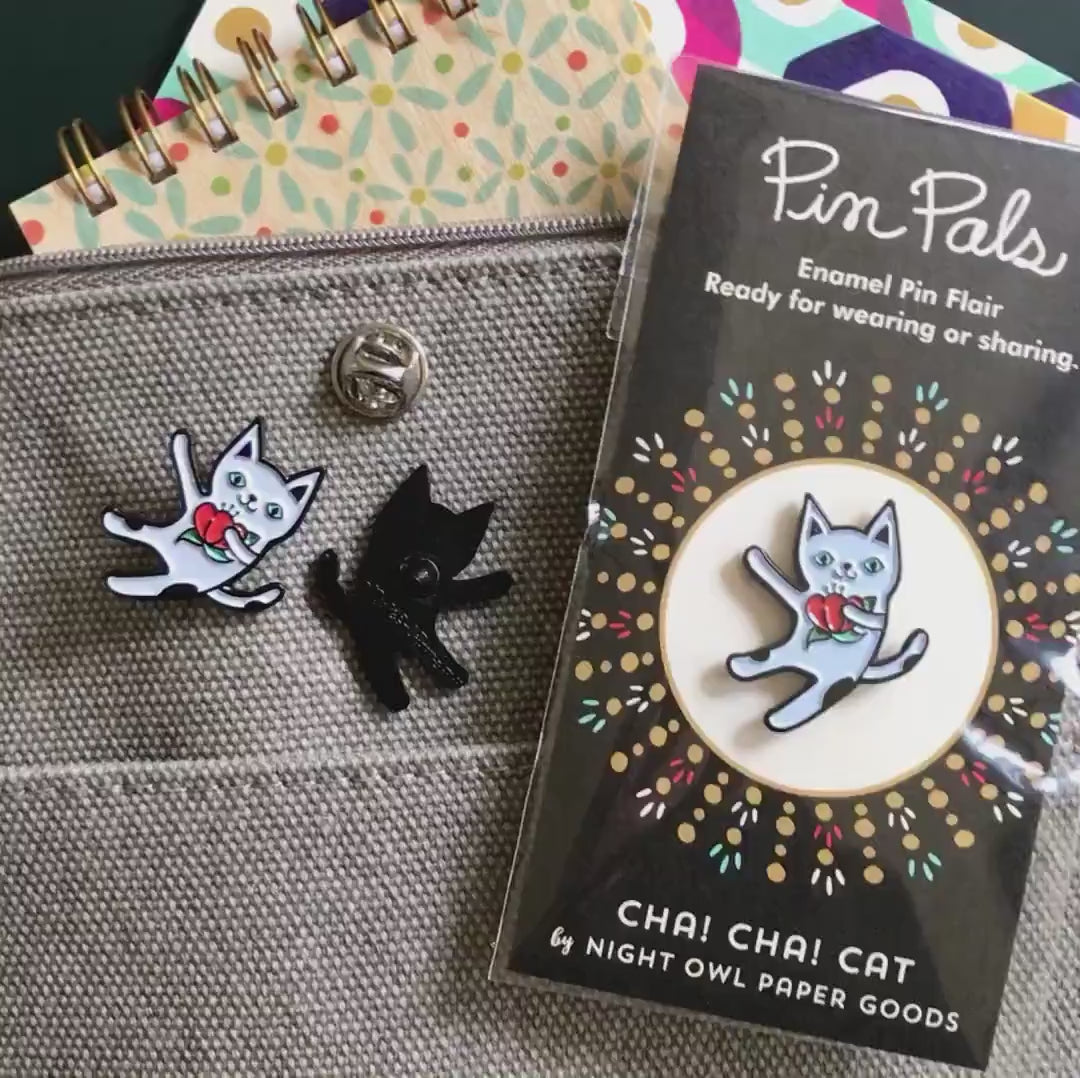 Front of enamel pin is a dancing white cat with black spots holding a red flower with black accents. Back of pin is black with the Night Owl Paper Goods script logo. Pin is mounted on a backer card that says "Pin Pals - Enamel Pin Flair - Ready for wearing or sharing - CHA! CHA! CAT - by Night Owl Paper Goods" and is packaged in a clear bag.