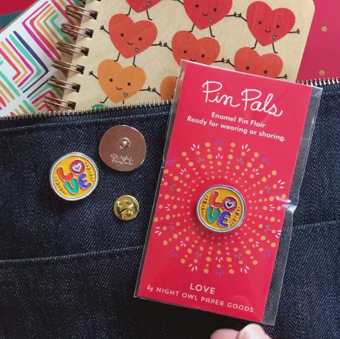 Front of enamel pin is yellow with colorful LOVE text, a small heart in the middle of the "O" and gold accents. Back of pin is gold with the Night Owl Paper Goods script logo. Pin is mounted on a backer card that says "Pin Pals - Enamel Pin Flair - Ready for wearing or sharing - LOVE - by Night Owl Paper Goods" and is packaged in a clear bag.