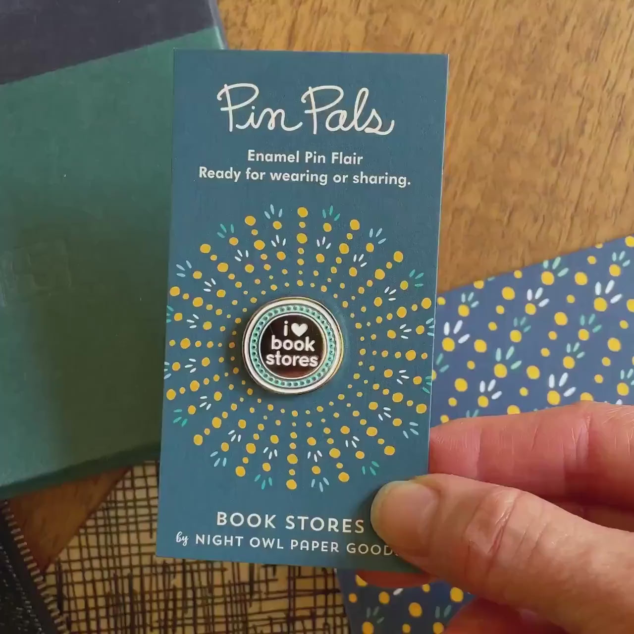Front of enamel pin is round with "I heart book stores" text and gold accents. Back of pin is gold with the Night Owl Paper Goods script logo. Pin is mounted on a backer card that says "Pin Pals - Enamel Pin Flair - Ready for wearing or sharing - BOOK STORES - by Night Owl Paper Goods" and is packaged in a clear bag.