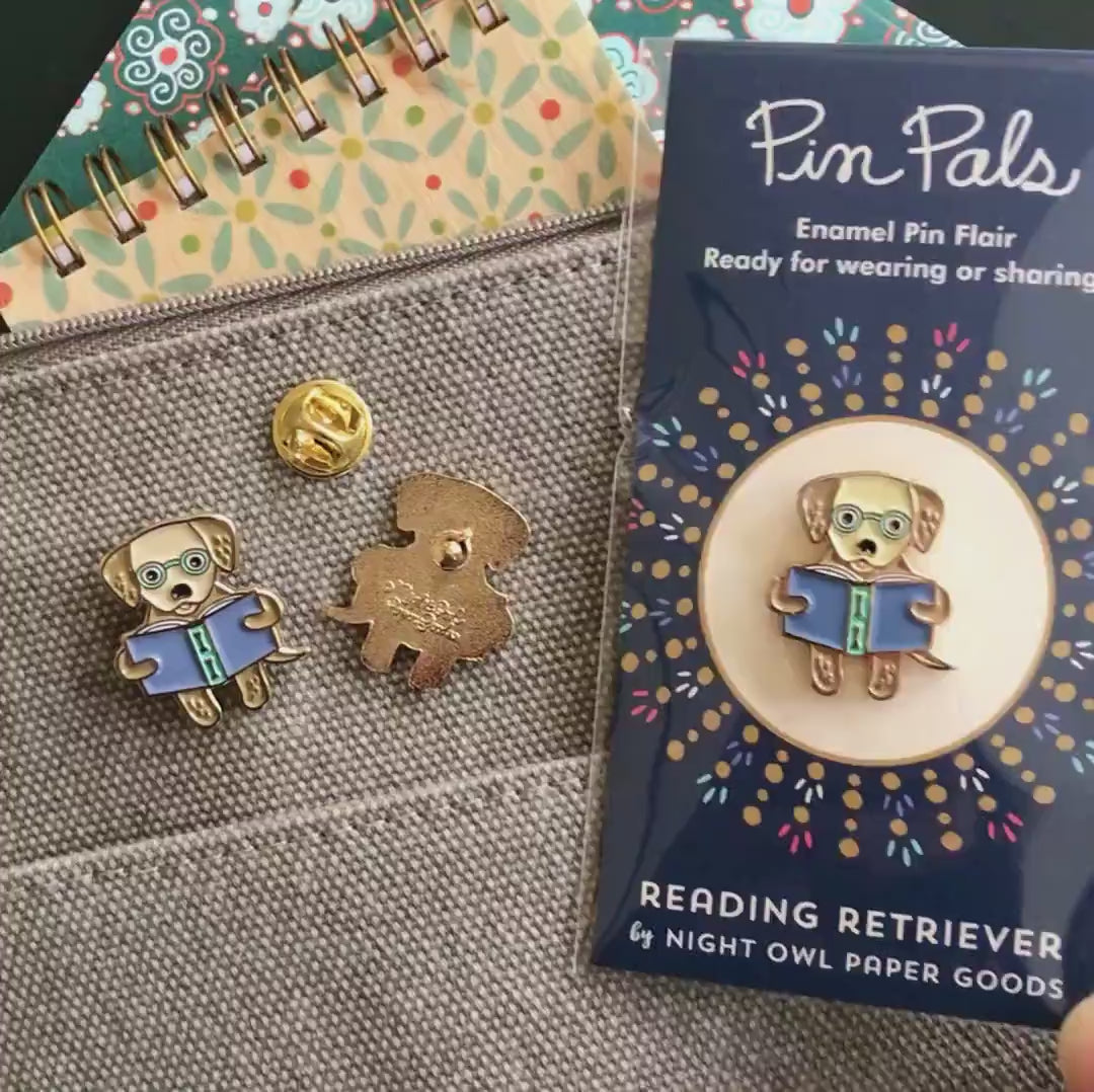 Front of enamel pin is a golden retriever reading a book with gold accents. Back of pin is gold with the Night Owl Paper Goods script logo. Pin is mounted on a backer card that says "Pin Pals - Enamel Pin Flair - Ready for wearing or sharing - READING RETRIEVER - by Night Owl Paper Goods" and is packaged in a clear bag.