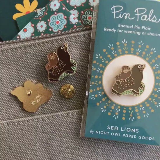 Front of enamel pin is a small sea lion on top of a larger sea lion with gold accents. Back of pin is gold with the Night Owl Paper Goods script logo. Pin is mounted on a backer card that says "Pin Pals - Enamel Pin Flair - Ready for wearing or sharing - SEA LIONS - by Night Owl Paper Goods" and is packaged in a clear bag.