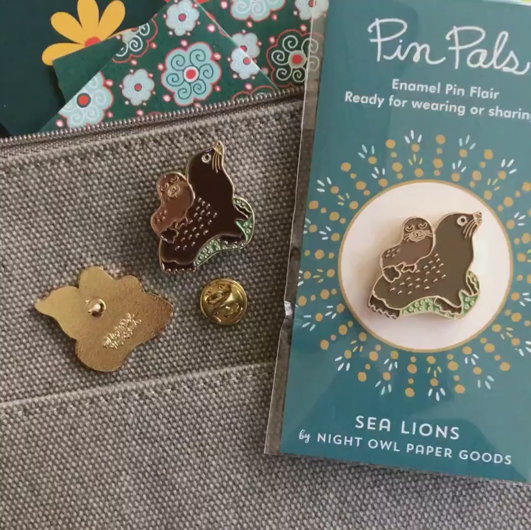 Front of enamel pin is a small sea lion on top of a larger sea lion with gold accents. Back of pin is gold with the Night Owl Paper Goods script logo. Pin is mounted on a backer card that says "Pin Pals - Enamel Pin Flair - Ready for wearing or sharing - SEA LIONS - by Night Owl Paper Goods" and is packaged in a clear bag.
