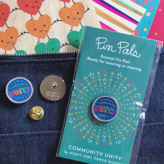 Front of enamel pin is blue with gold COMMUNITY text, rainbow UNITY text and gold accents. Back of pin is gold with the Night Owl Paper Goods script logo. Pin is mounted on a backer card that says "Pin Pals - Enamel Pin Flair - Ready for wearing or sharing - COMMUNITY UNITY - by Night Owl Paper Goods" and is packaged in a clear bag.