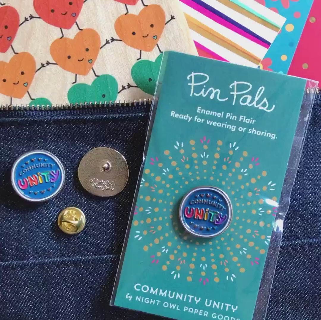 Front of enamel pin is blue with gold COMMUNITY text, rainbow UNITY text and gold accents. Back of pin is gold with the Night Owl Paper Goods script logo. Pin is mounted on a backer card that says "Pin Pals - Enamel Pin Flair - Ready for wearing or sharing - COMMUNITY UNITY - by Night Owl Paper Goods" and is packaged in a clear bag.