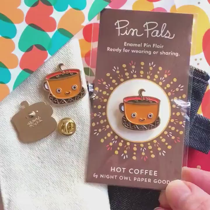 Front of enamel pin is a smiling coffee mug with gold accents. Back of pin is gold with the Night Owl Paper Goods script logo. Pin is mounted on a backer card that says "Pin Pals - Enamel Pin Flair - Ready for wearing or sharing - HOT COFFEE - by Night Owl Paper Goods" and is packaged in a clear bag.