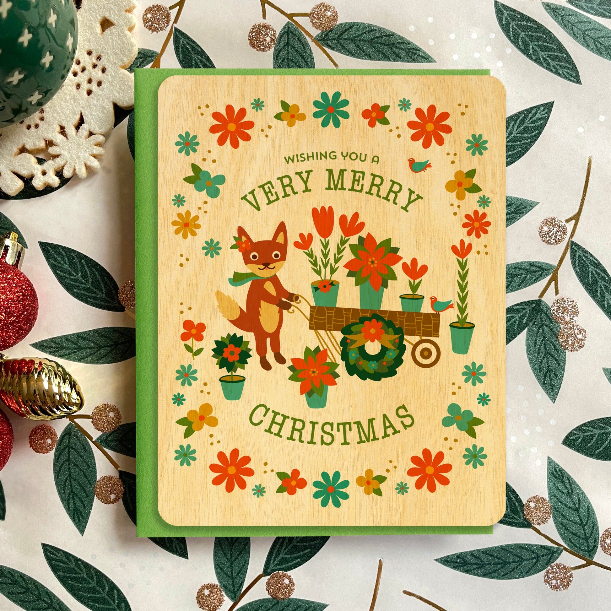 Thin wood christmas card with illustrated fox pushing a festive flower cart full of red flowers, wreaths, and poinsettias.