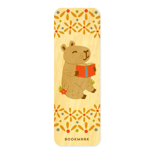 Front of Capy birch wood mini bookmark featuring a cute capybara holding an open red book, surrounded by decorative floral patterns in orange, yellow, green and blue at the top and bottom.